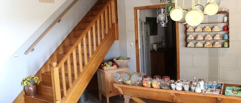 A wooden staircase leading to a kitchen with a wooden table and bench, a hanging pot rack, and a shelf with jars and kitchenware.