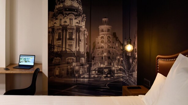 Habitación de hotel con cama, escritorio con computadora portátil, mesita de noche con lámpara y un gran mural de paisaje urbano en la pared.