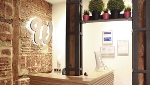 A reception desk with a wooden counter, a brick wall, and a modern light fixture. There are potted plants on a shelf above the desk and a Wi-Fi sign on the wall.