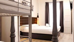 A hotel room with a bunk bed, a large bed, a bedside lamp, and a view of the outside through curtains.