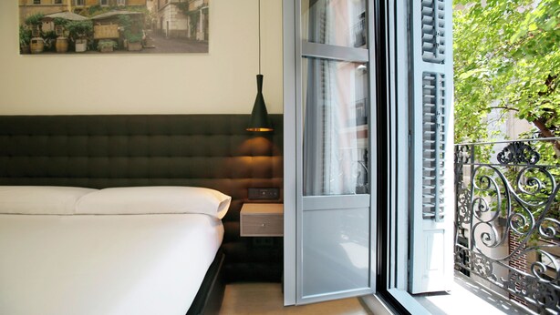 A bedroom with a bed, bedside table, lamp, and a view of a balcony with greenery.