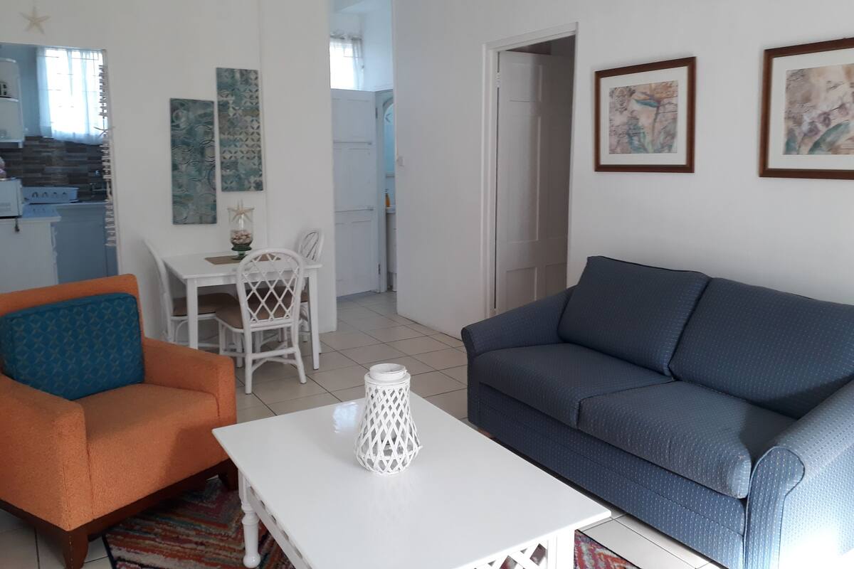 A living room with a blue sofa, orange armchair, and a white coffee table. There is a dining area with a white table and chairs.