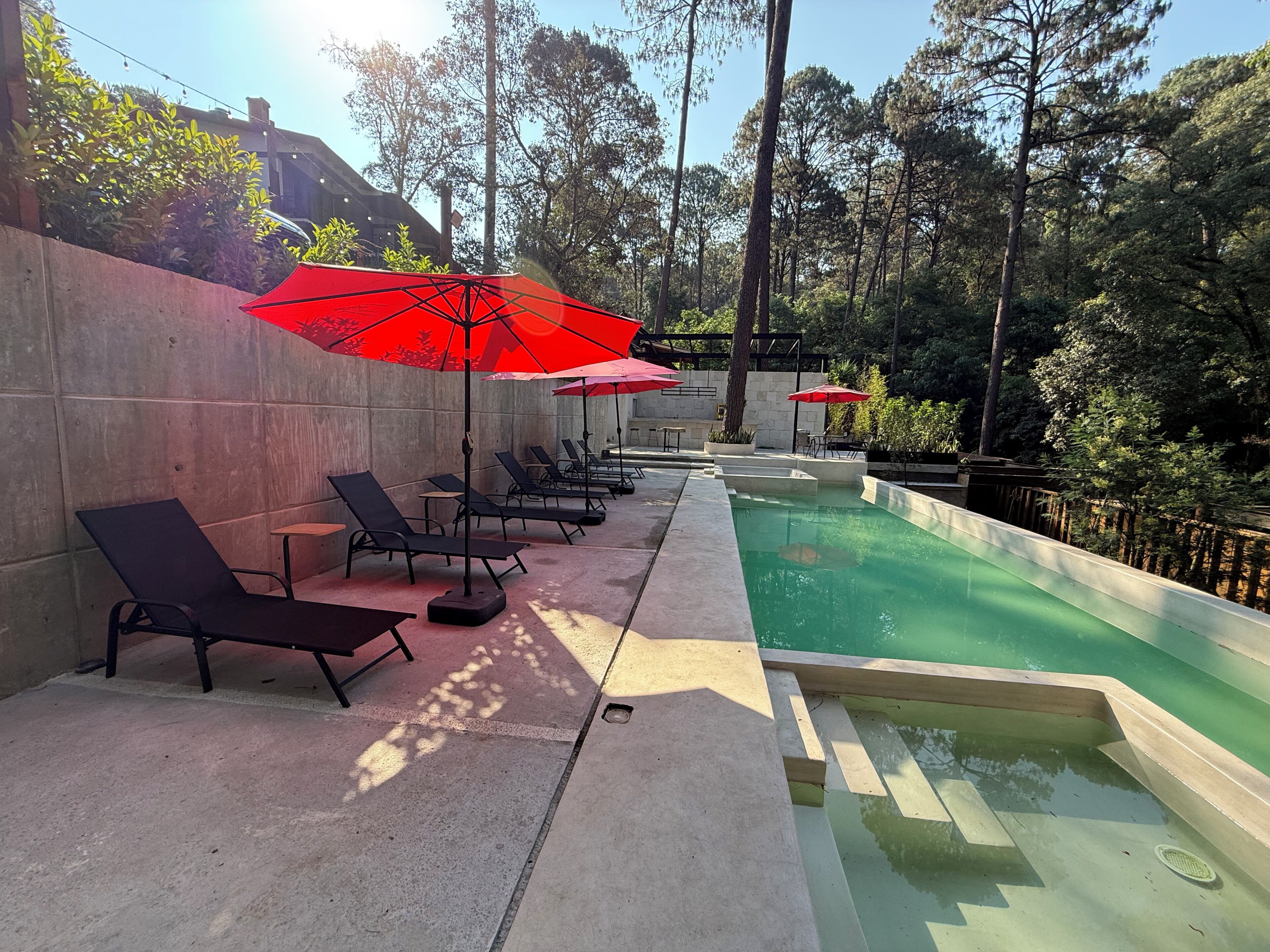 A poolside area with lounge chairs and red umbrellas, surrounded by trees.
