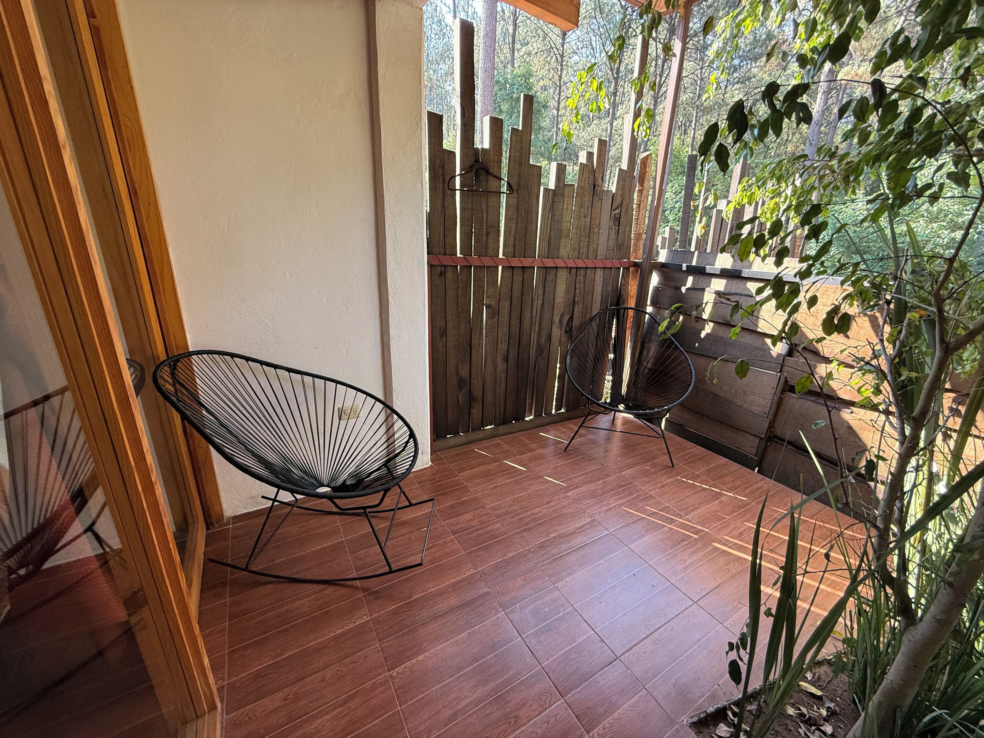 A wooden-floored balcony with two black wire-framed chairs, a wooden fence, and a view of greenery.