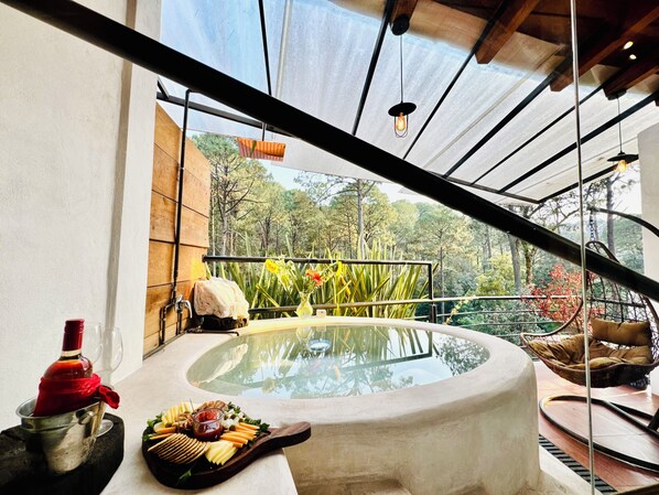 Una mesa con cubierta de vidrio, varios alimentos y una botella, frente a un balcón con vistas a árboles y una lámpara colgante.