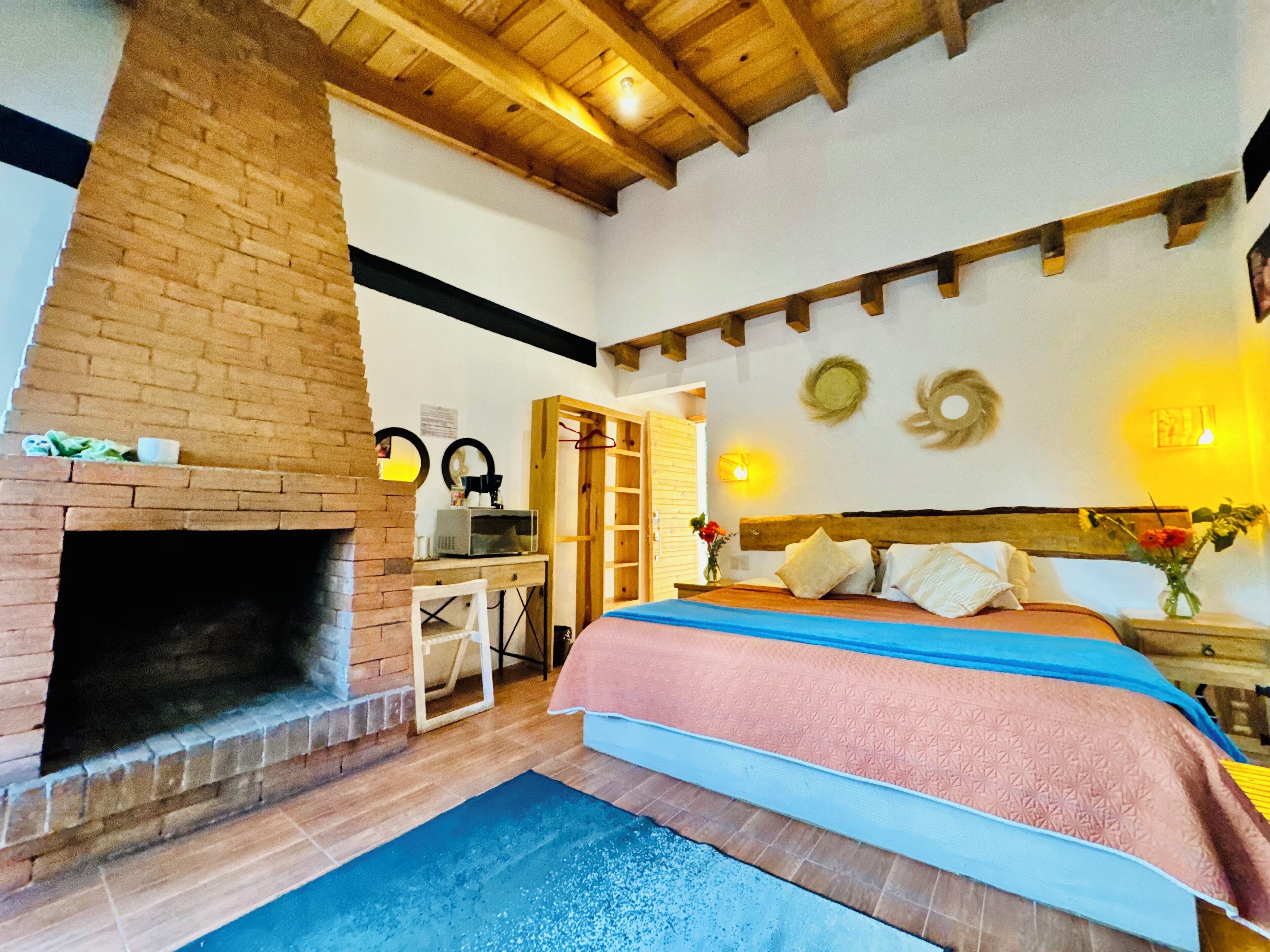 A bedroom with a brick fireplace, a bed with a blue and pink bedspread, a desk with a computer, and a wooden ceiling.