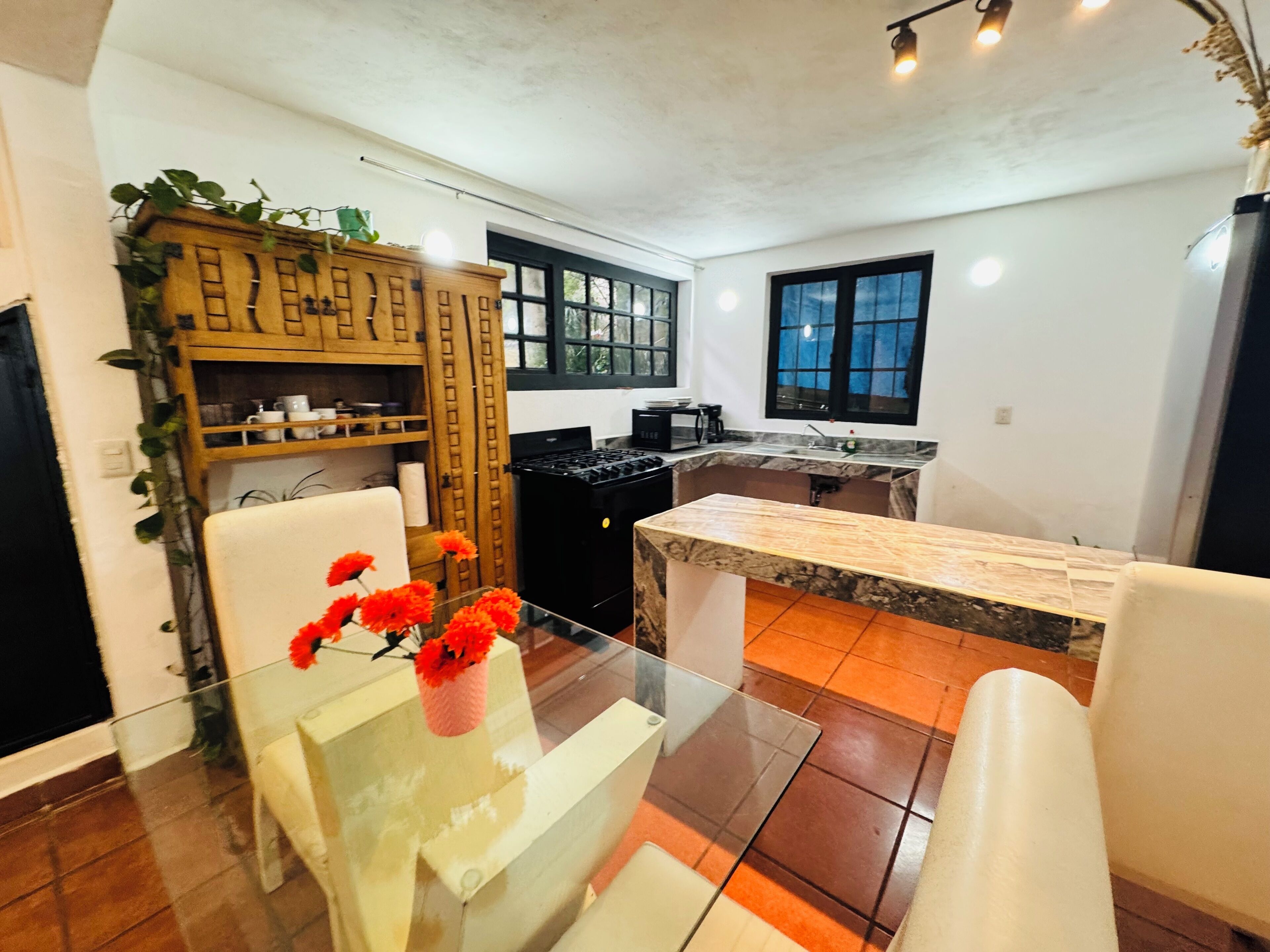 A modern kitchen with a marble countertop, a glass dining table with a vase of flowers, and a wooden cabinet with shelves.