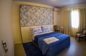 Ein Schlafzimmer mit einem blumengemusterten Kopfteil, einem Bett mit blauer Bettdecke, einem Sessel und einem Fenster mit Vorhängen.