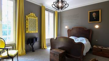 A bedroom with a wooden bed, a small wooden chest, a chair, a radiator, and a window with yellow curtains.