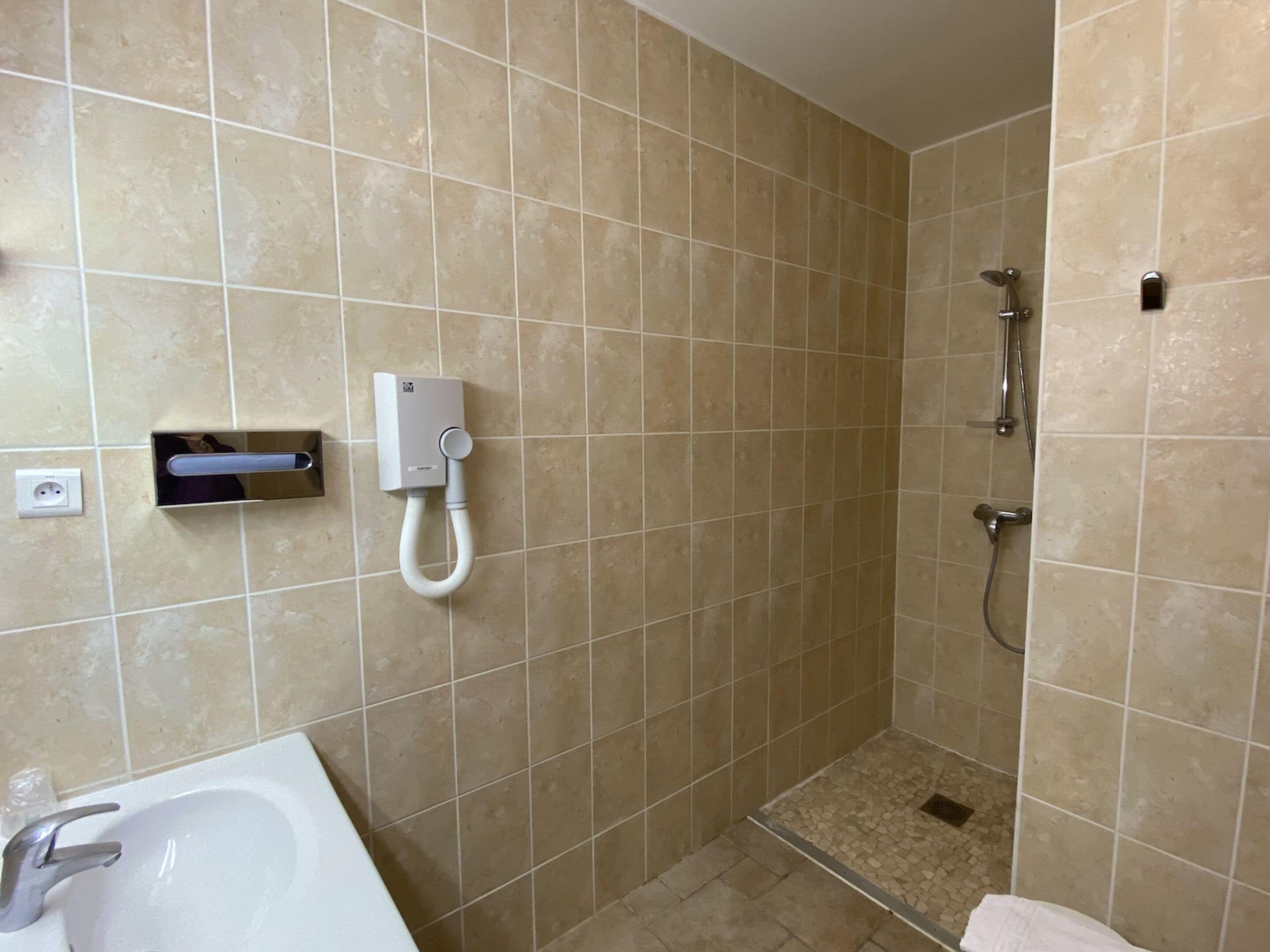 Une salle de bain avec des carreaux de couleur beige, un lavabo blanc et une douche équipée d’un pommeau de douche à main.