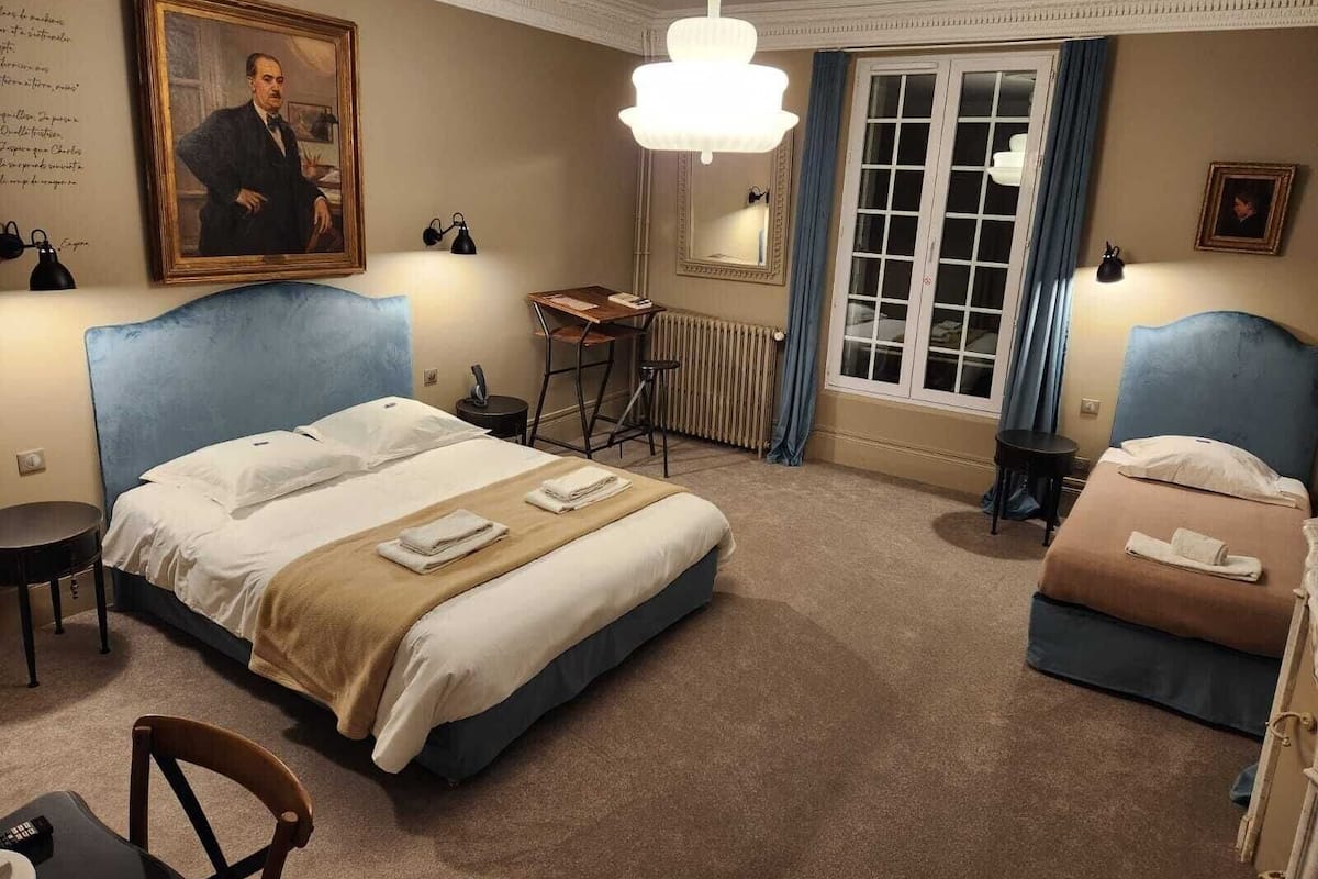 A hotel room with a large bed, a smaller bed, a desk, a chair, a radiator, a window with curtains, and a painting on the wall.