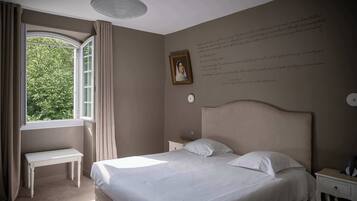 Une chambre à coucher avec un grand lit, une table de chevet, une commode, une fenêtre avec des rideaux et un mur orné d’un tableau encadré et d’un texte.