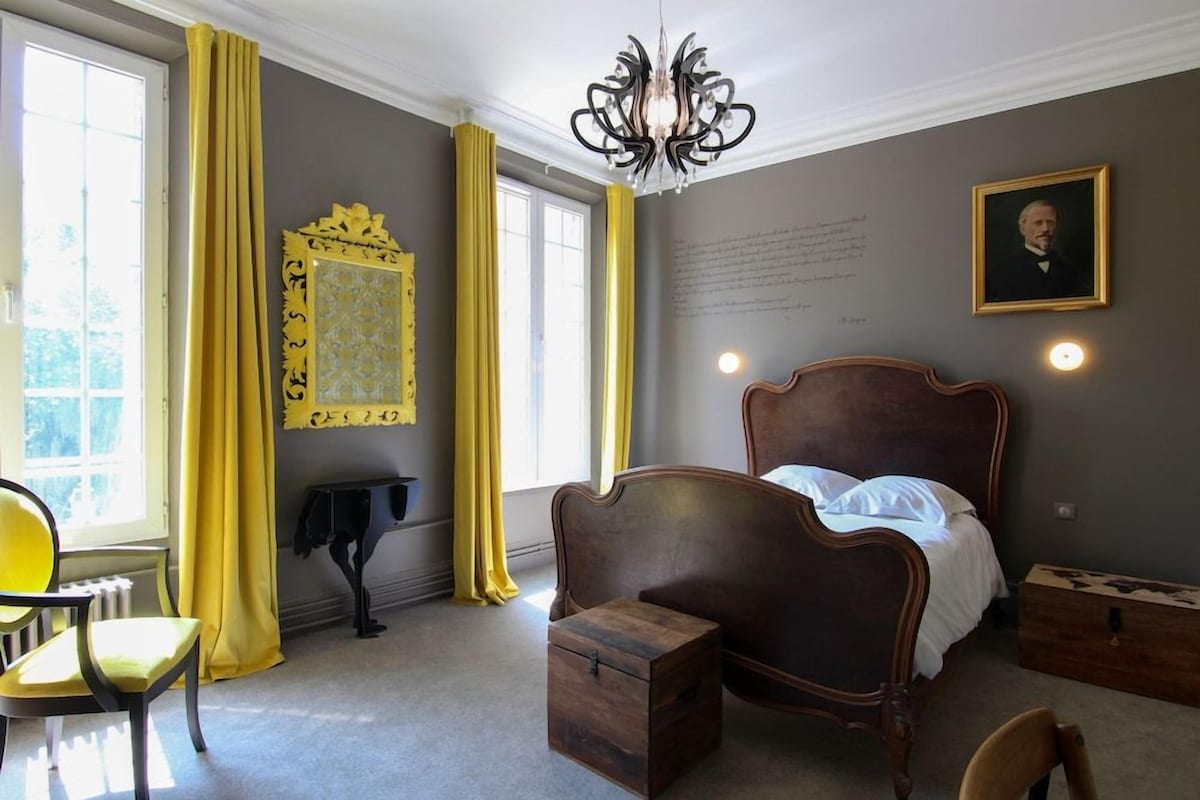 Una habitación con una cama grande, una silla, una mesita, un espejo y una araña.