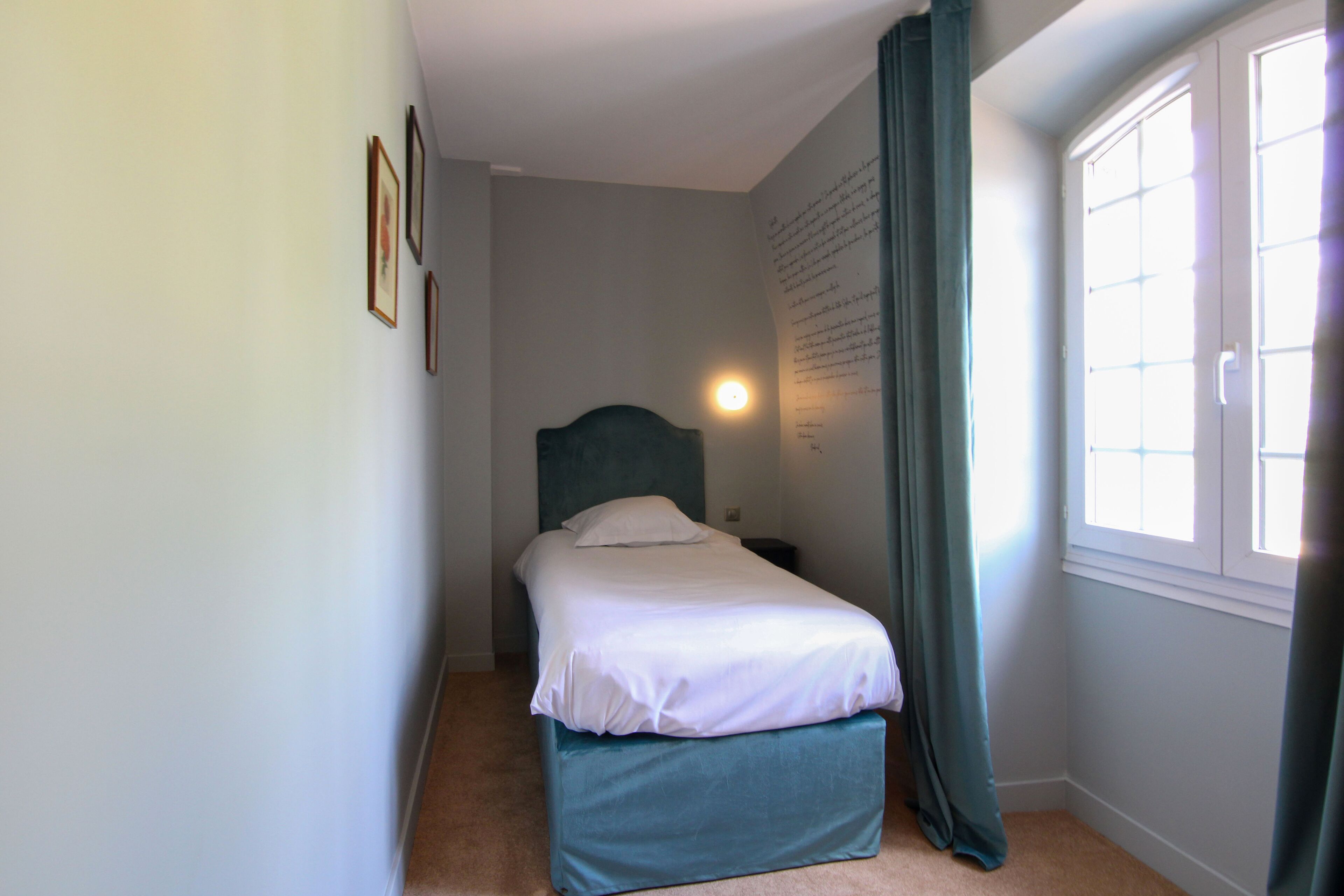 A small, neatly arranged bedroom with a single bed, a nightstand, a wall-mounted light, and a window with curtains.