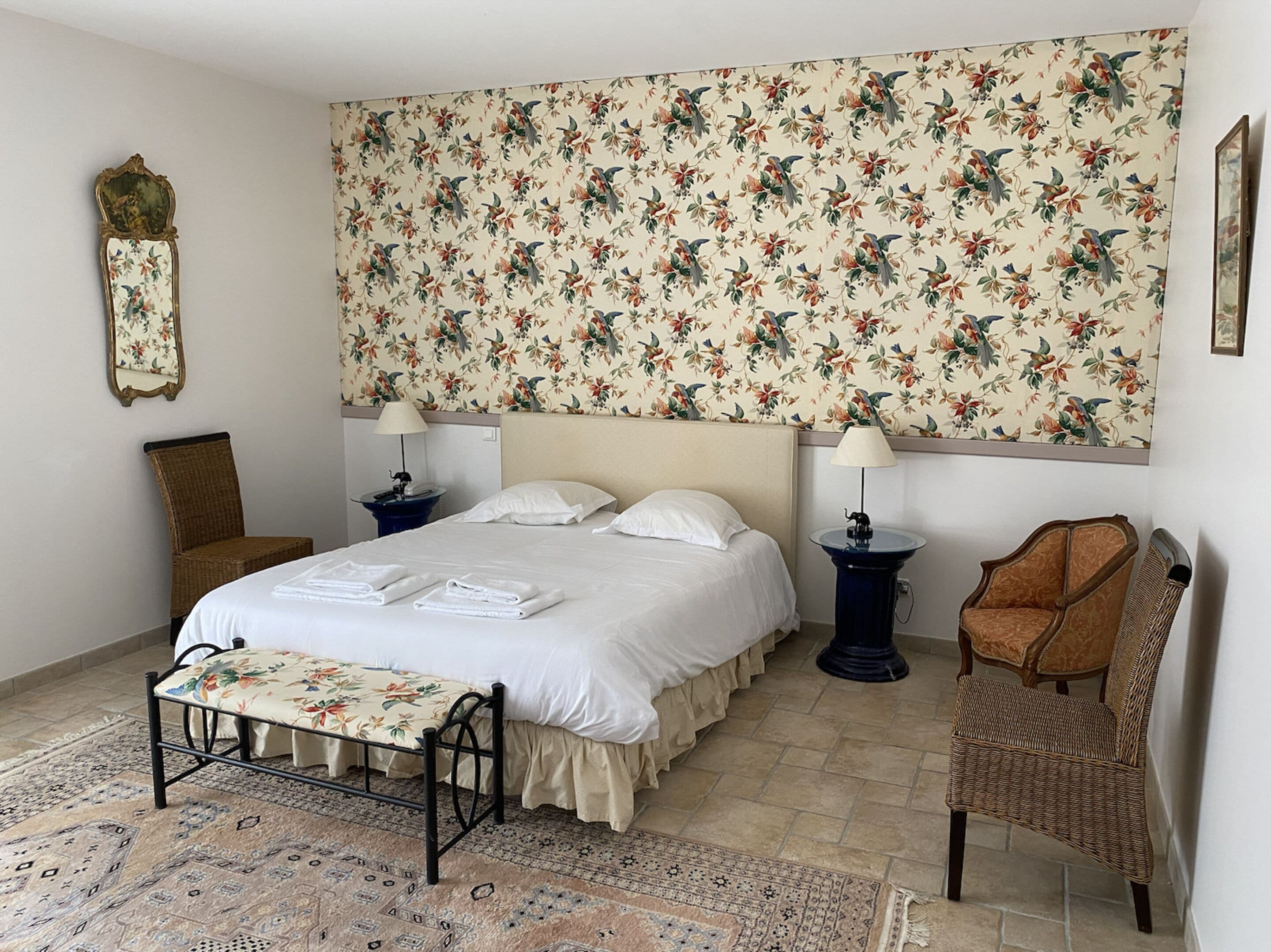 Ein ordentlich eingerichtetes Schlafzimmer mit einem Bett, zwei Nachttischen, einem Sessel und einer Bank, versehen mit einer auffälligen floralen Tapete.