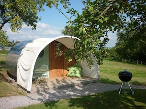 Une petite structure semi-ouverte, semblable à une tente, avec une table et des chaises à l’intérieur, un coussin vert à l’extérieur et un barbecue à proximité.