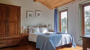 Quarto com guarda-roupa de madeira, cama com cobertor azul, duas luminárias de mesa e janela com cortinas.