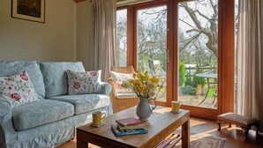 Uma sala de estar aconchegante com um sofá azul claro, uma mesa de centro de madeira com livros e um vaso de flores, e uma grande janela com vista para um jardim e móveis de exterior.