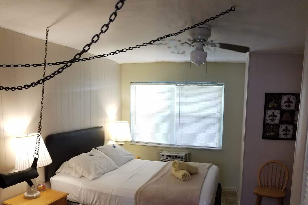 Une chambre d’hôtel avec un lit, des lampes de chevet, un ventilateur de plafond, une fenêtre avec des stores et une chaise.