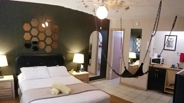 Habitación de hotel con una cama grande, lámparas de noche, una silla colgante, una kitchenette pequeña y un espejo en la pared.
