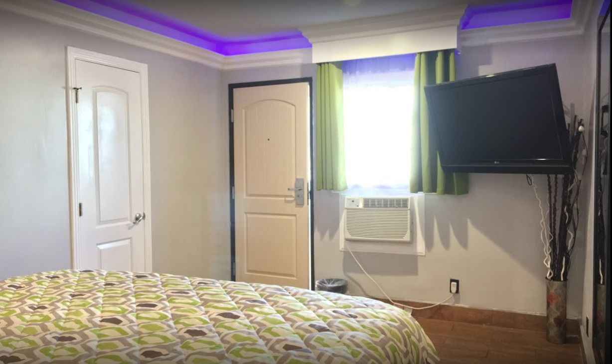 A bedroom with a patterned bedspread, a closed door, an air conditioning unit, a television mounted on the wall, and a window with curtains.