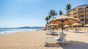 Ein Strand mit Liegestühlen und Sonnenschirmen, ein Gebäude mit Balkonen und Palmen.