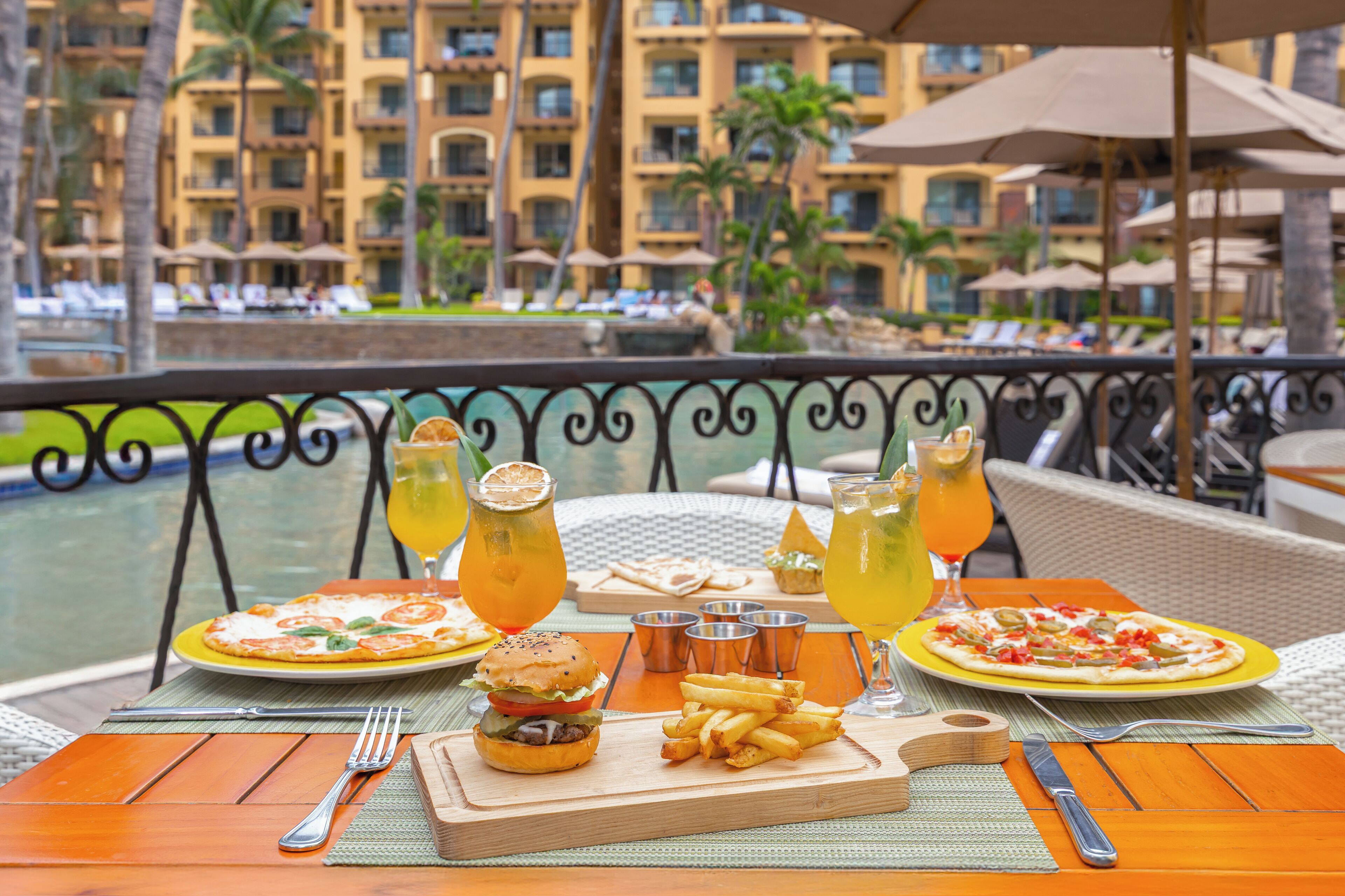 Una mesa de comedor con una hamburguesa, papas fritas y pizza, junto con bebidas, frente a una piscina de hotel y palmeras.