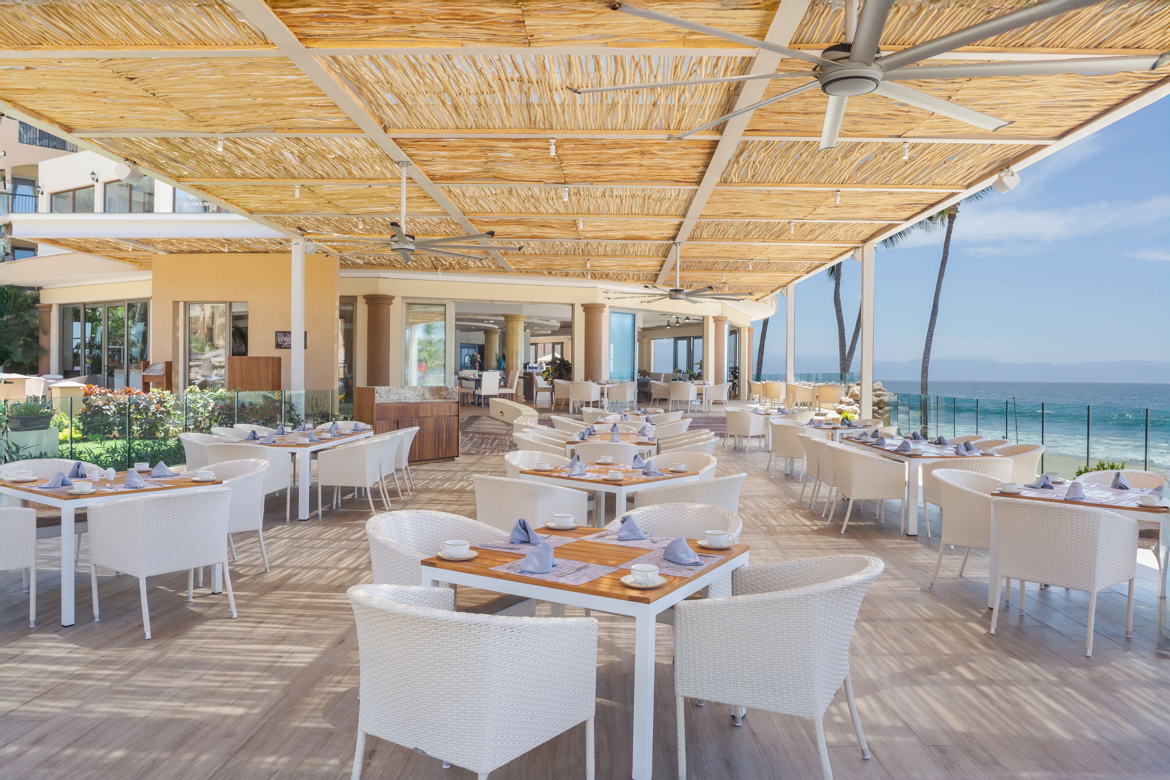 An outdoor dining area with white wicker furniture, a thatched roof, and a view of the ocean.