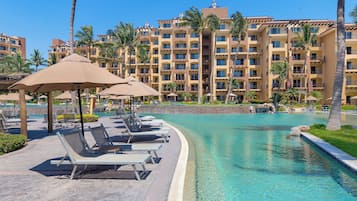 A resort pool area with lounge chairs and umbrellas, surrounded by palm trees and a large hotel building in the background.
