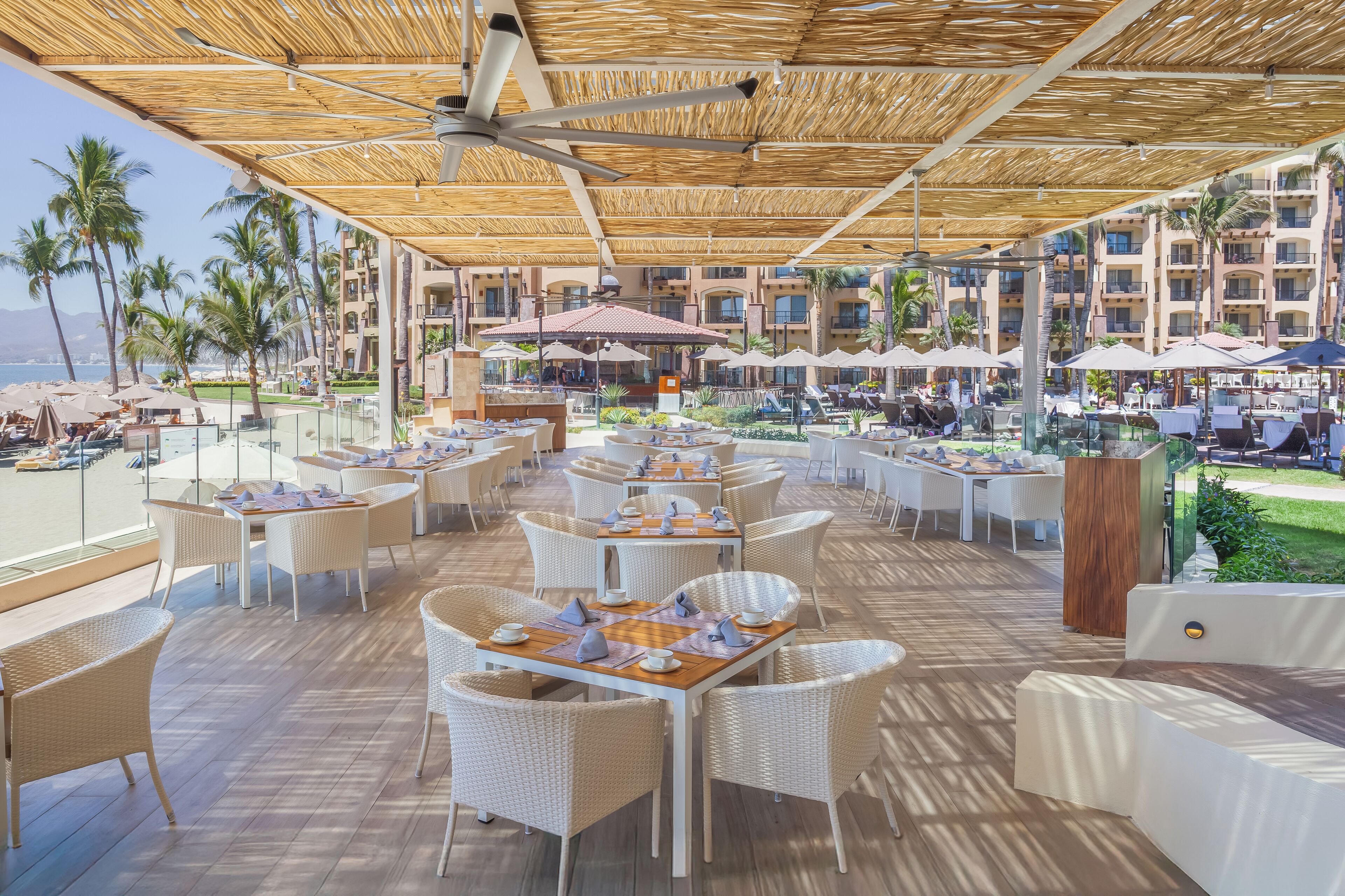 Un espacio exterior cubierto para comer con mobiliario de mimbre y sombrillas, con vistas a una piscina y palmeras.