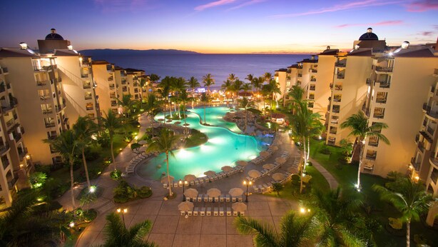 Ein Resort mit einem zentralen Pool, Palmen und Blick auf den Ozean bei Sonnenuntergang.
