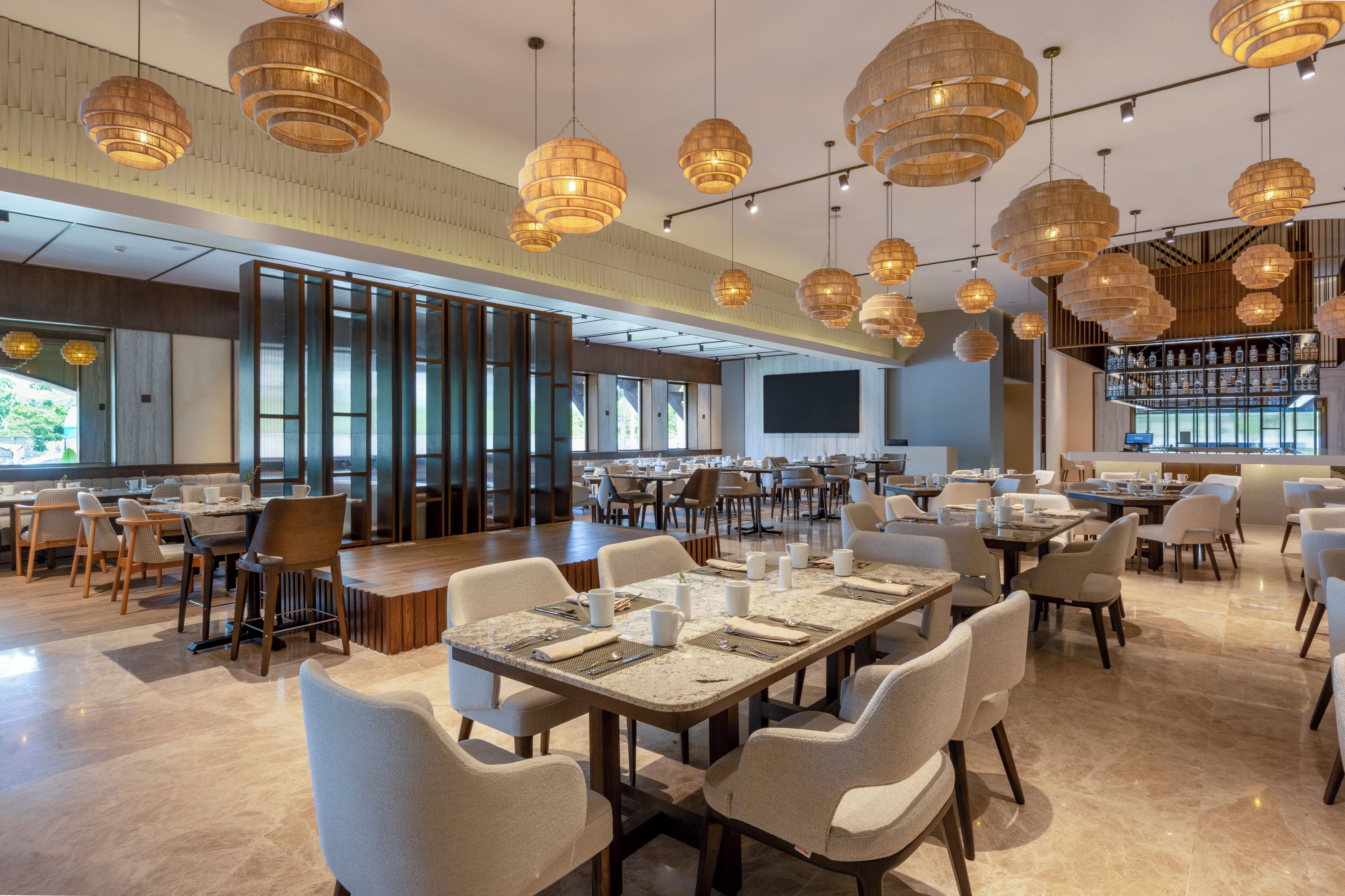 A spacious restaurant with multiple dining tables and chairs, large windows, and hanging wooden pendant lights.