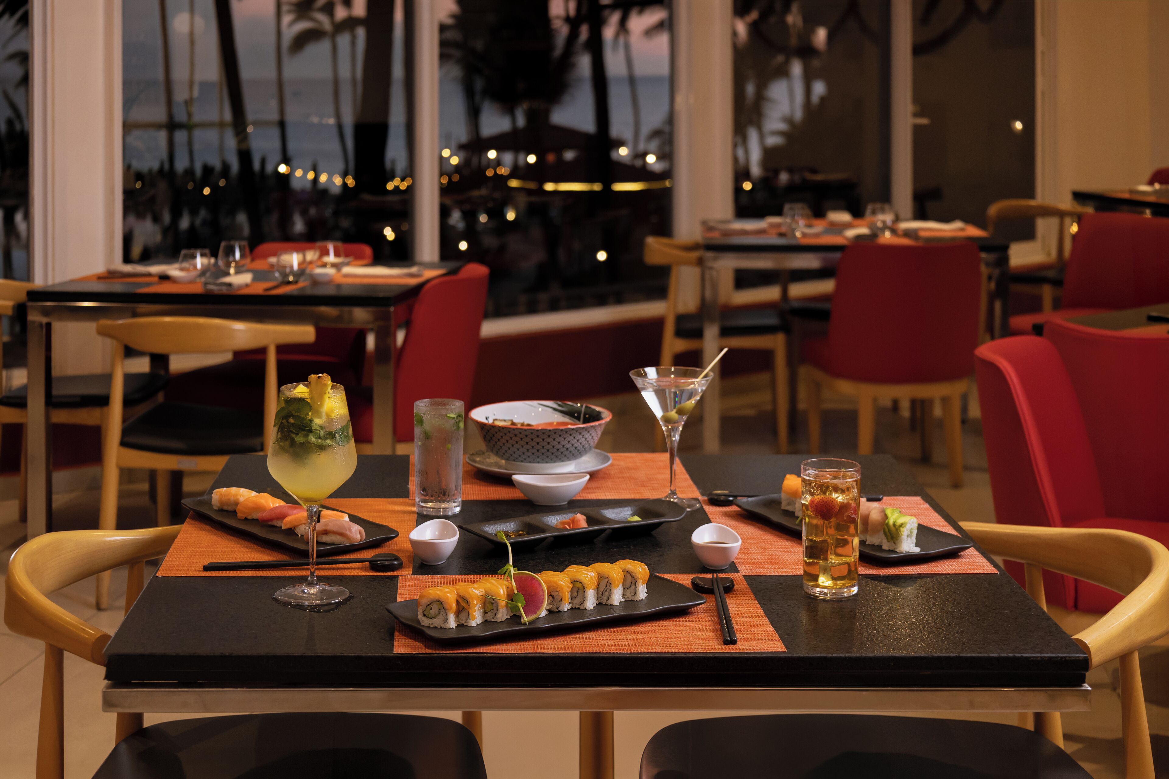 A restaurant with a view of palm trees and a beach, sushi dishes, and a martini glass on the table.