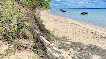 A sandy beach with boats in the water, greenery on the shore, and a clear sky.