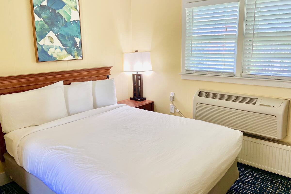 A hotel room with a bed, a nightstand, a lamp, a window with blinds, and a wall-mounted air conditioner.