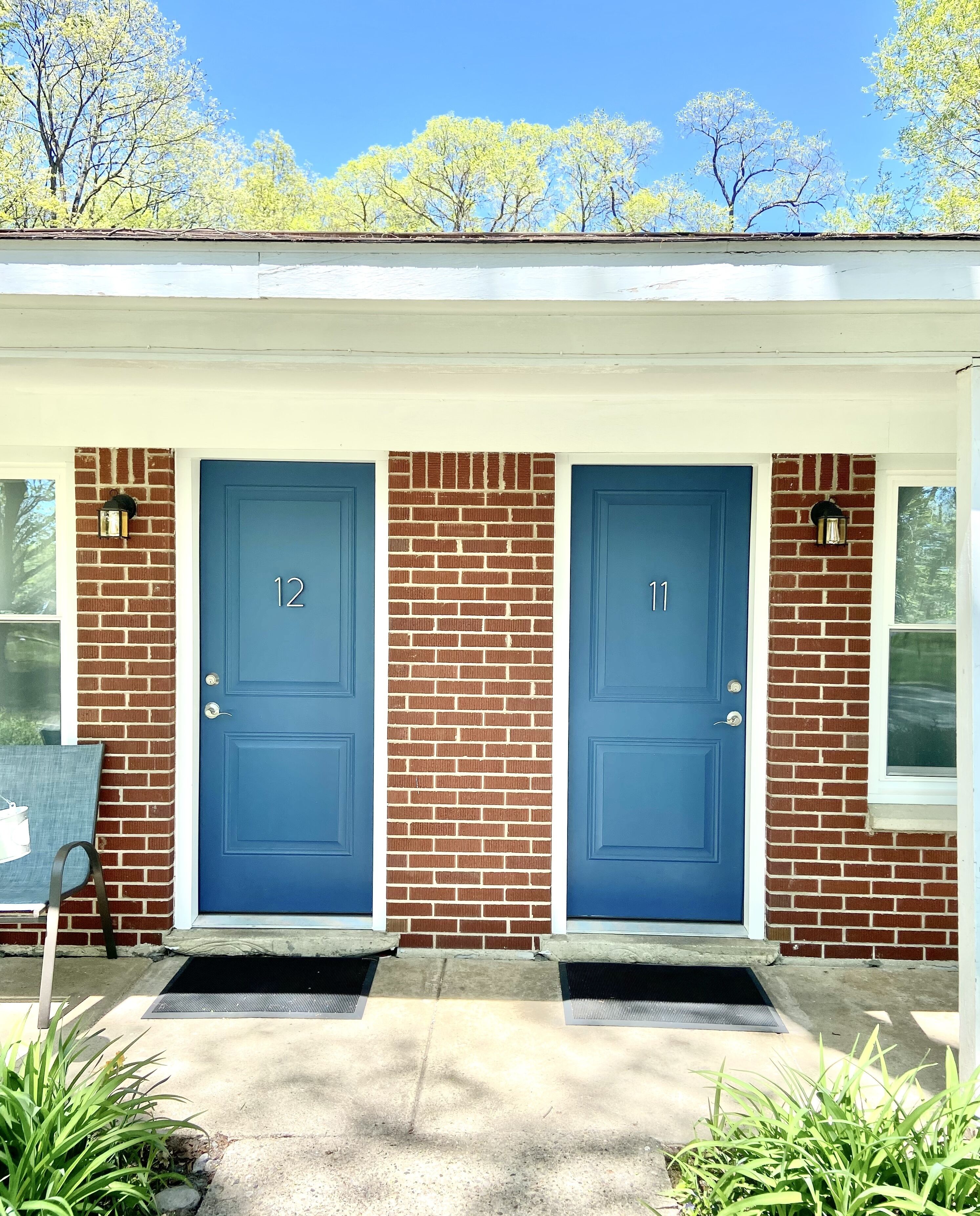 A two-story house with a brick facade, two blue doors labeled 12 and 11, and a small porch area with a bench and plants.