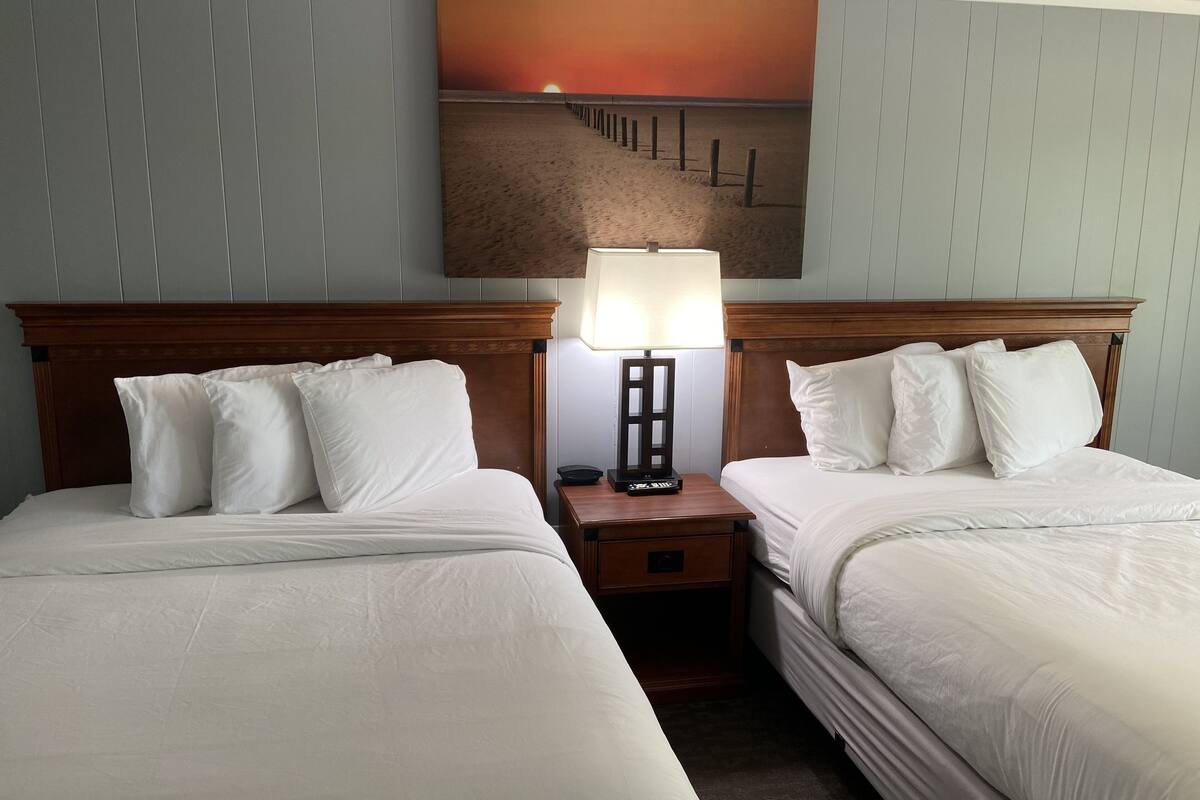 A hotel room with two beds, a nightstand with a lamp, and a painting of a sunset on the wall.