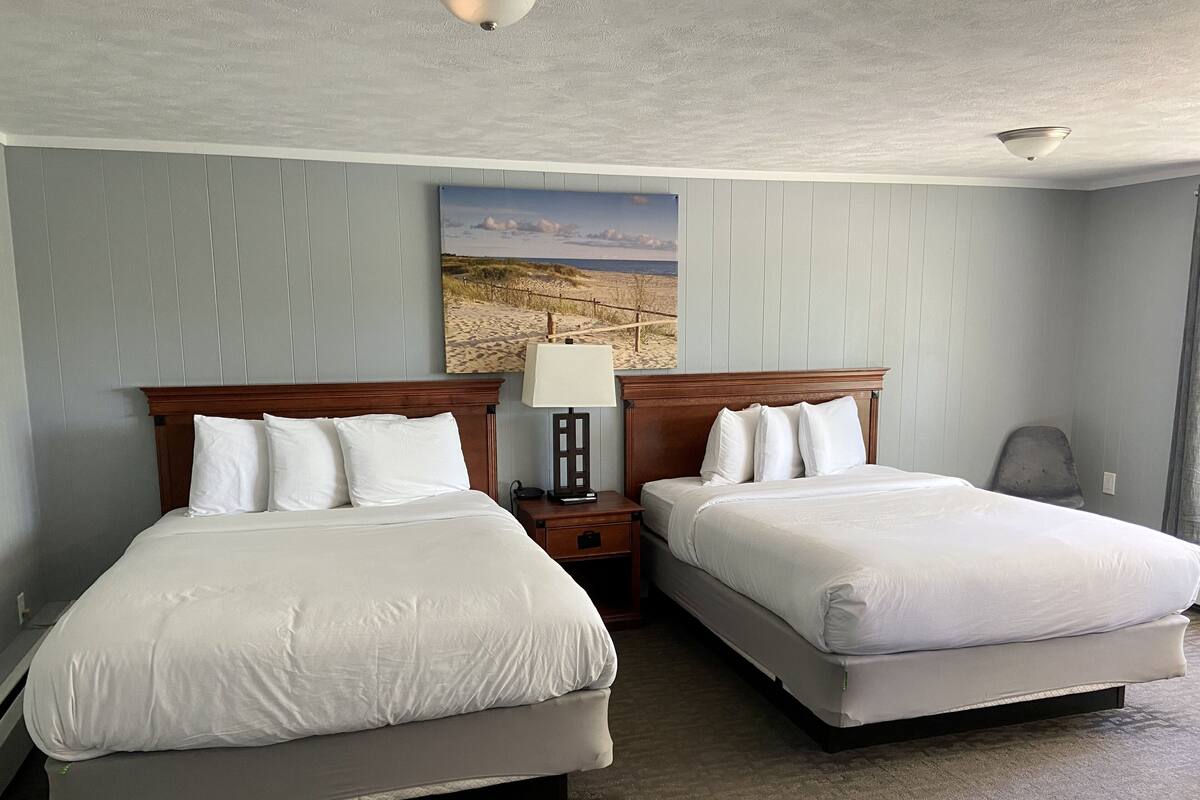 A hotel room with two beds, a nightstand, a lamp, and a painting of a beach scene.