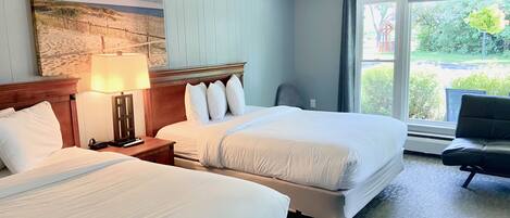 A hotel room with two beds, a nightstand with a lamp, a large window with a view of greenery, and a painting on the wall.