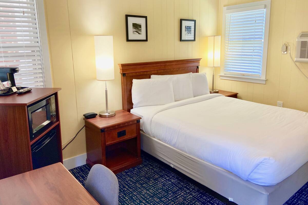 A hotel room with a large bed, bedside tables, a desk with a chair, and a window with blinds.