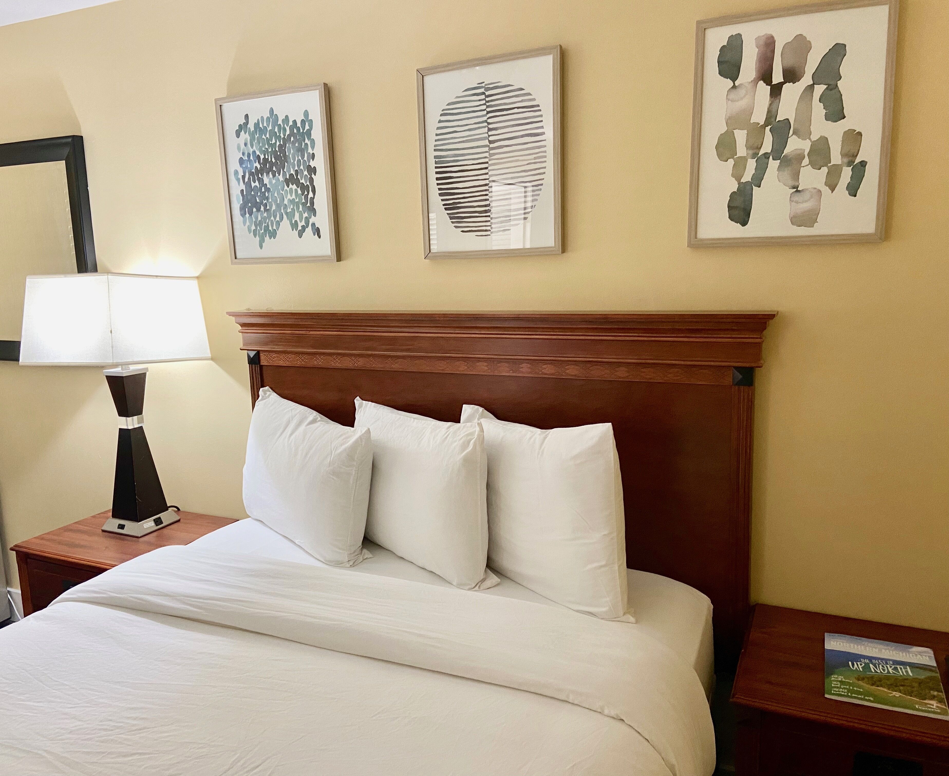 Una habitación de hotel con cabecera de madera, cama con ropa blanca, mesita de noche con lámpara y tres cuadros enmarcados en la pared.