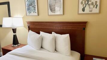 Una habitación de hotel con cabecera de madera, cama con ropa blanca, mesita de noche con lámpara y tres cuadros enmarcados en la pared.