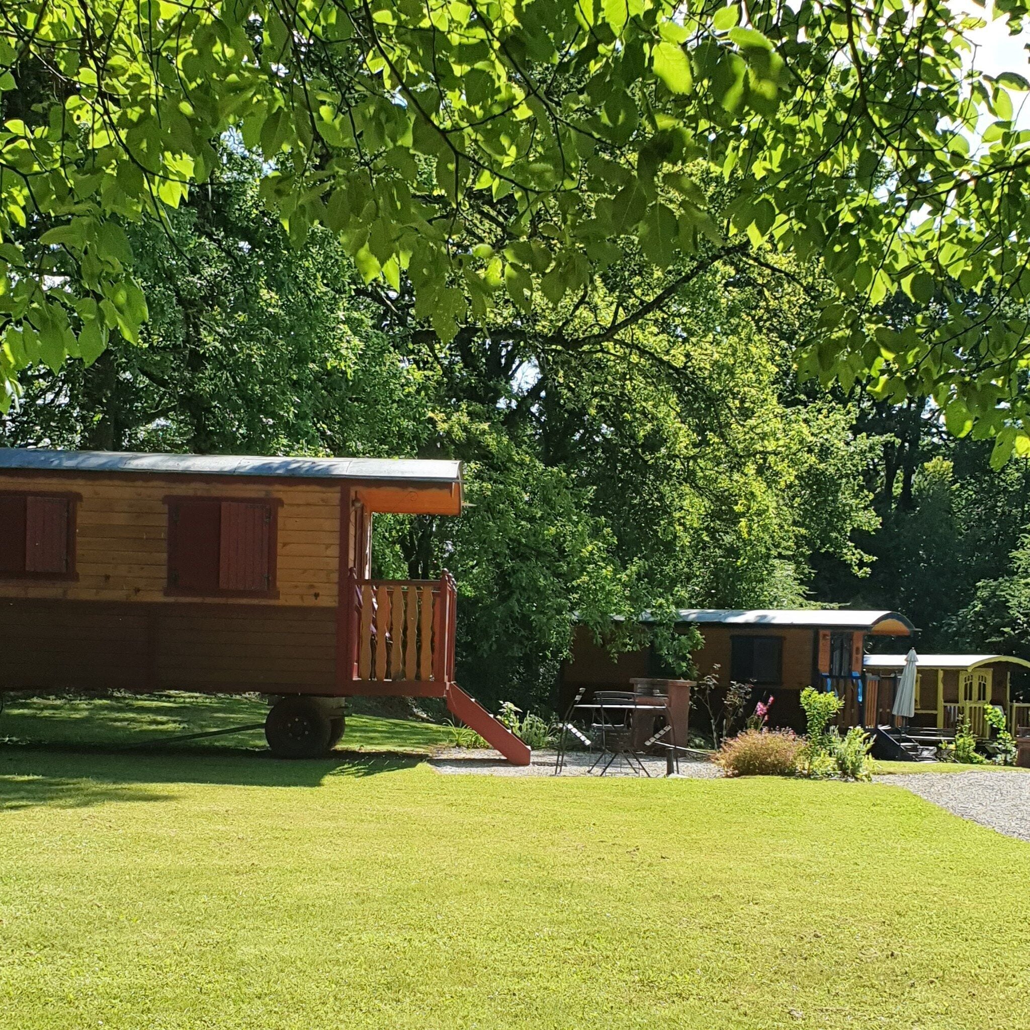 Un camper di legno con un portico e un piccolo tavolo, immerso in alberi verdi e rigogliosi.