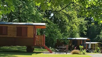 Un camper di legno con un portico e un piccolo tavolo, immerso in alberi verdi e rigogliosi.