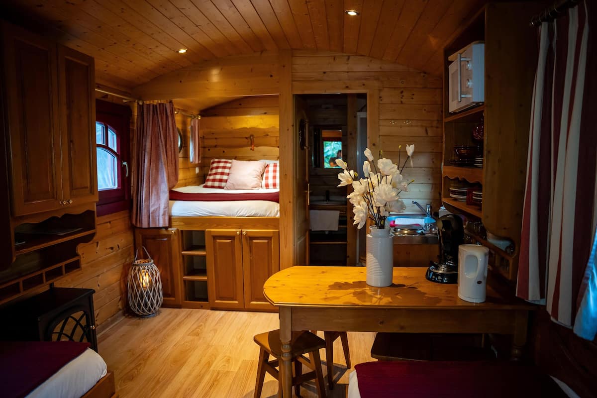 A cozy cabin interior with a wooden interior, a bed with red and white bedding, a dining table with chairs, and a kitchen area with a kettle and flowers.