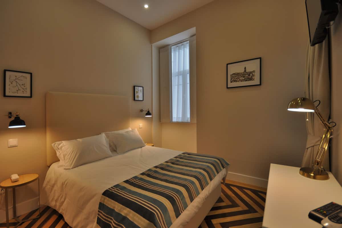 Una habitación de hotel con cama, mesita de noche, lámpara y una ventana con cortinas.
