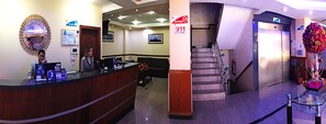 A hotel reception area with a reception desk, a 'No Fumes' sign, an 911 emergency sign, a staircase, and a floral arrangement. - Hotel Novalux (Ambato)