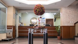 A hotel reception area with a reception desk, a large floral arrangement, a potted plant, and a luggage cart. - Hotel Novalux (Ambato)