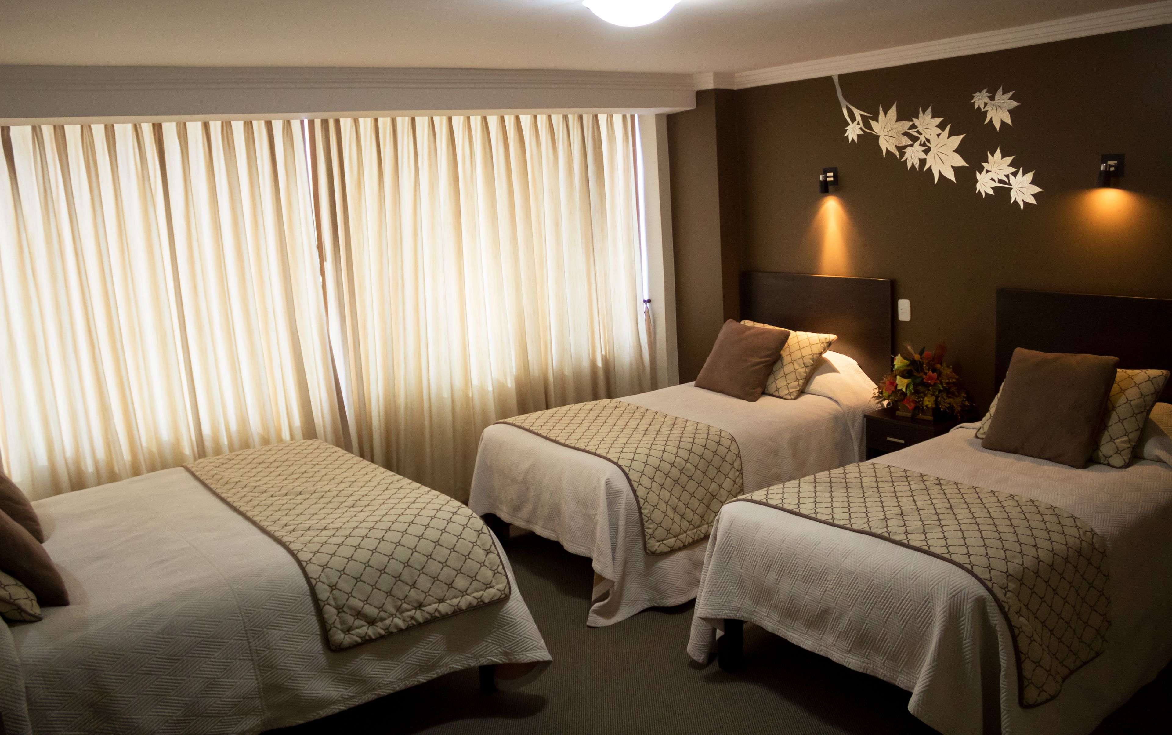 A hotel room with two single beds, a large window with curtains, a wall-mounted light fixture, and a decorative leaf pattern on the wall.
