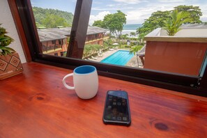 A wooden table with a white mug and a smartphone. Through a window, a pool and a building with a red roof are visible. - Mar Arena 27 Apartment (Jacó)